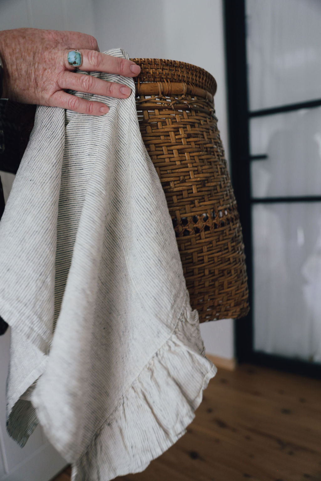 Hand holding a Saltbush living pinstripe french linen Lisiére tea towel with a cane basket 