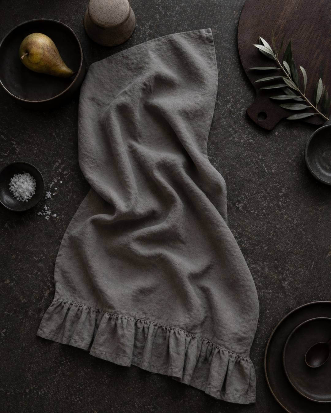 Saltbush Living french linen Lisiére Tea Towel with a frill in slate colour lying on a kitchen bench next to some salt, a pear and a small olive branch of leaves