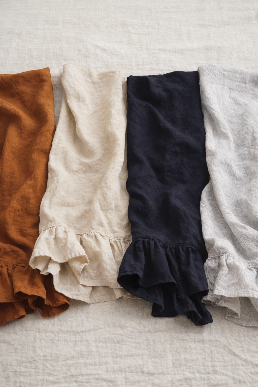 4 different coloured Saltbush Living french linen Lisiére Tea Towels with a frill in rust, black, natural and pinstripe lying on a large piece of linen