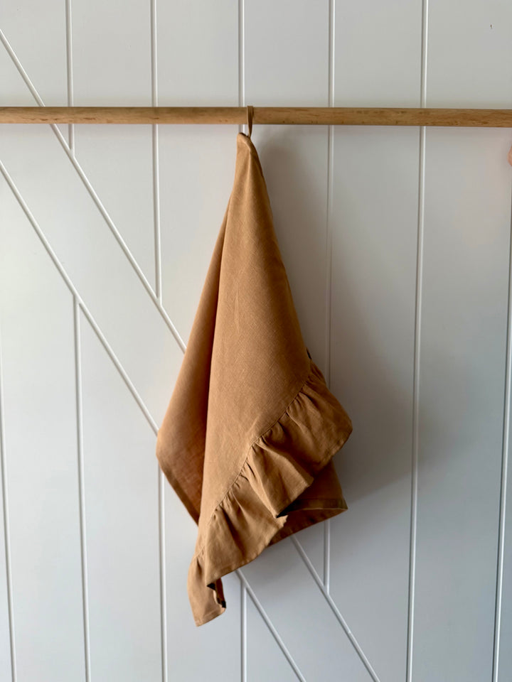 Toffee coloured Lisiére linen tea towel hanging from a wooden dowel against a textured white wall.