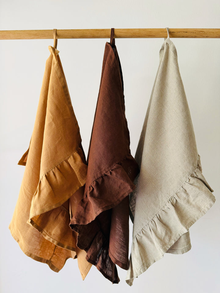 Three textured Lisiére linen tea towels in toffee, chocolate, and lnatural, hanging from a wooden dowel against a plain white wall.
