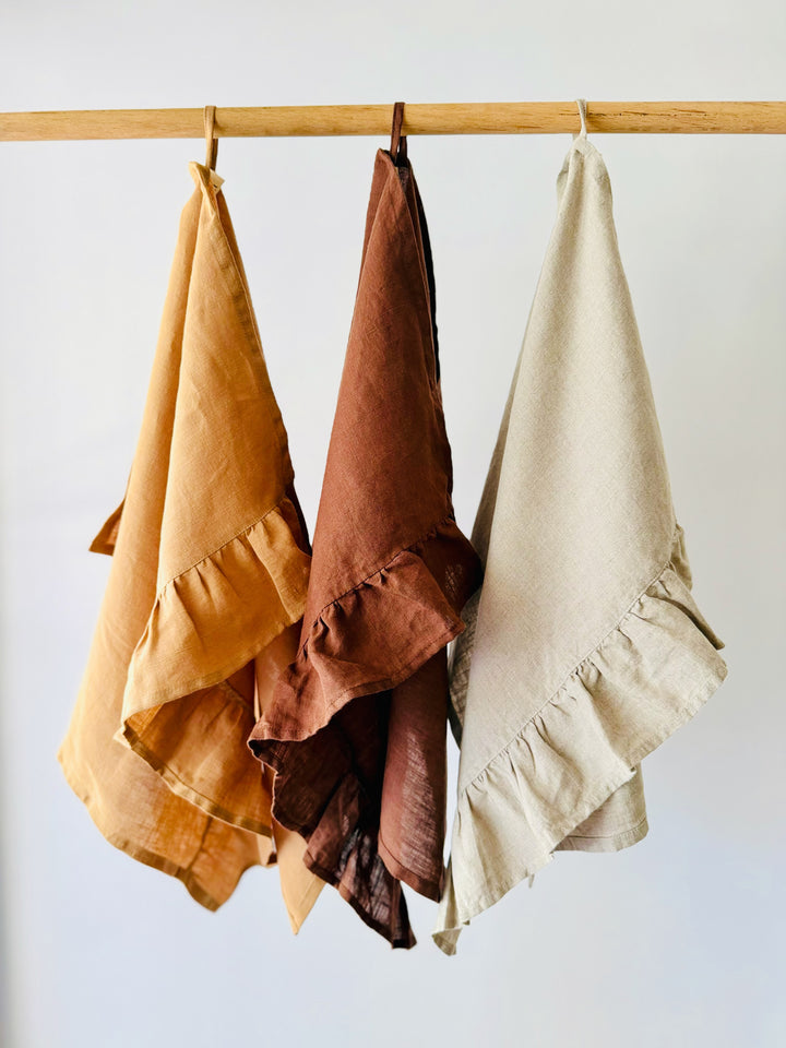 Three textured Lisiére linen tea towels in toffee, chocolate, and lnatural, hanging from a wooden dowel against a plain white wall.
