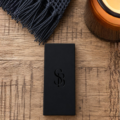 A black rectangular matchbox with Saltbush Living embossed monogram logo, placed on a rustic wooden surface next to a dark woven textile and part of an amber candle jar.