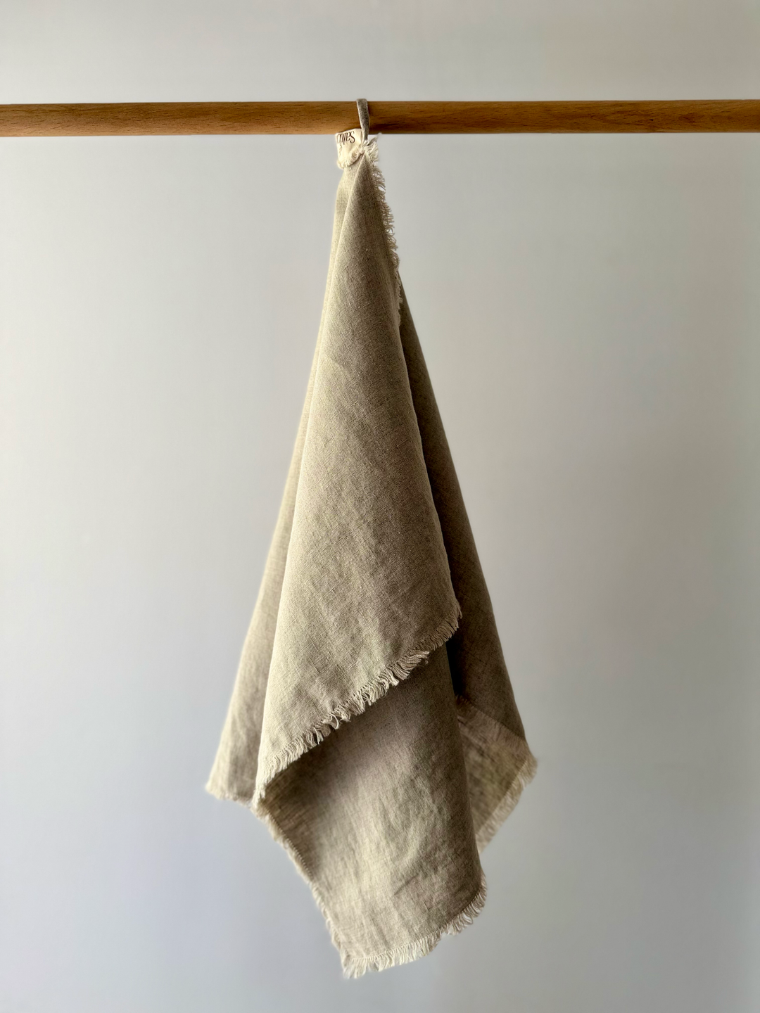 A natural linen tea towel with frayed edges hangs from a linen loop on a wooden rod against a plain light gray wall.